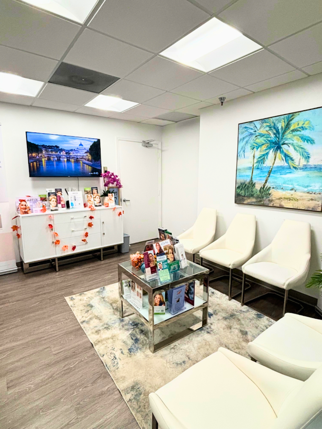 Modern waiting room with comfortable white chairs, glass coffee table, large TV, and beautiful palm tree artwork creating a welcoming coastal atmosphere